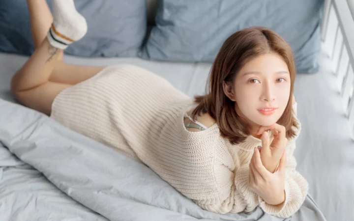 一位成年女性穿着米白色针织裙趴在床上，面向镜头微笑。