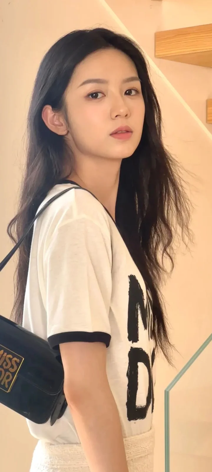 一名长发女性在室内回头面对镜头，身穿白色短袖上衣，背景是暖色墙面和木质装饰。