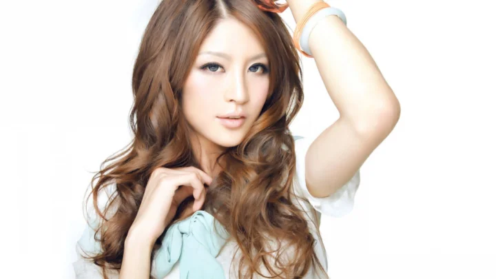 長い髪の女性が白い背景の前でポートレートのポーズを取っている。