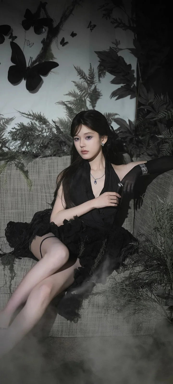 一名身穿黑色服装的女性坐在蕨类植物和深色叶片环绕的室内背景前。