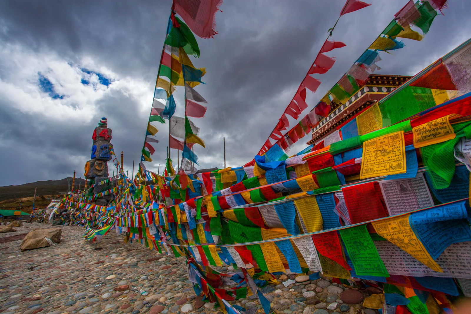 五彩经幡在乌云密布的高原天空下飘扬，远处可见山地与石地。