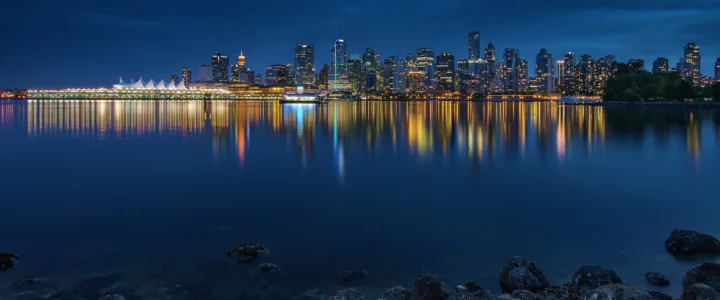 夜色中的城市天际线倒映在平静的水面上，前景可见岸边岩石。