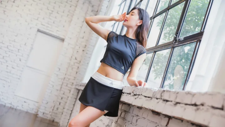 黒いスポーツウェアを着た成人女性が、明るい窓と白いレンガ壁のそばでポーズを取っている。