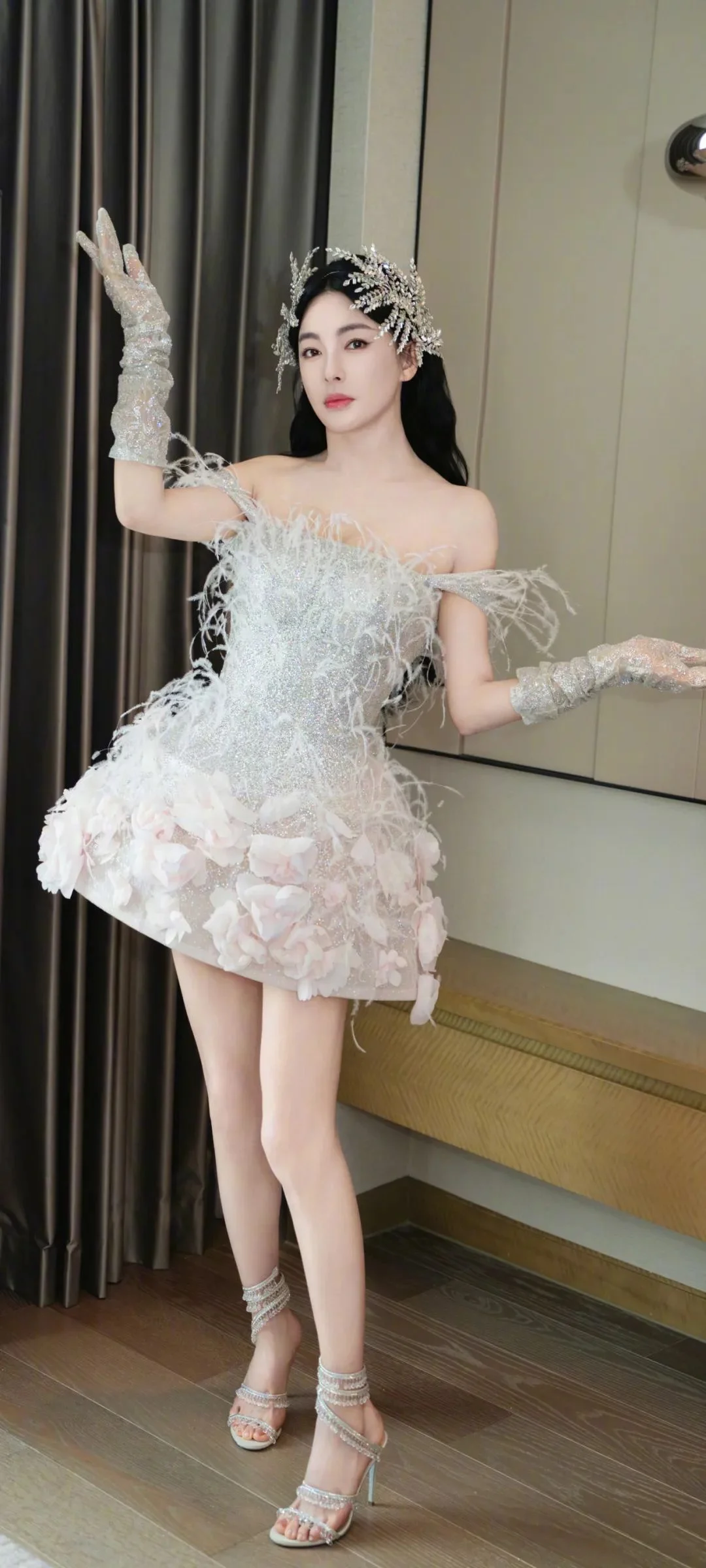白い羽根飾りのドレスとティアラ、長い手袋を身に着けた成人女性が室内のカーテンのそばに立っています。