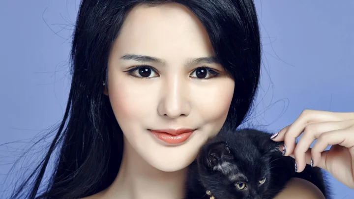 一名黑发女性面对镜头微笑，怀中抱着一只黑猫，背景为淡蓝色。