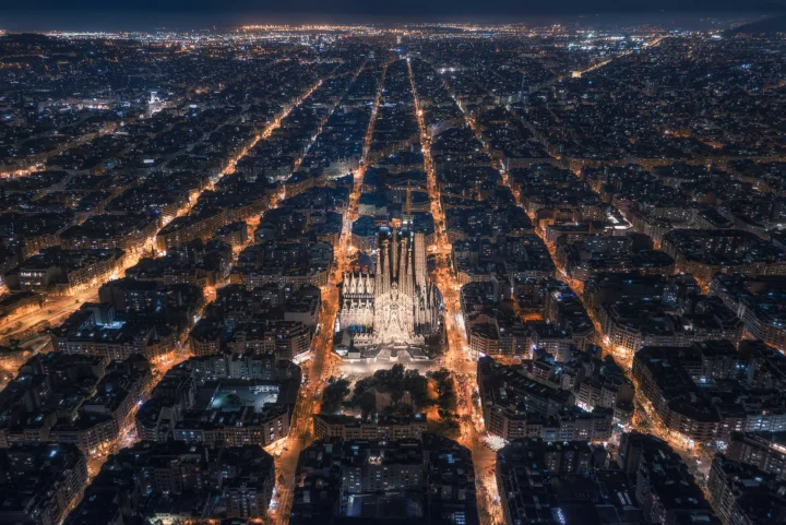 夜幕下的城市从高空俯瞰，中央一座灯火通明的尖顶建筑被纵横交错的街道环绕。