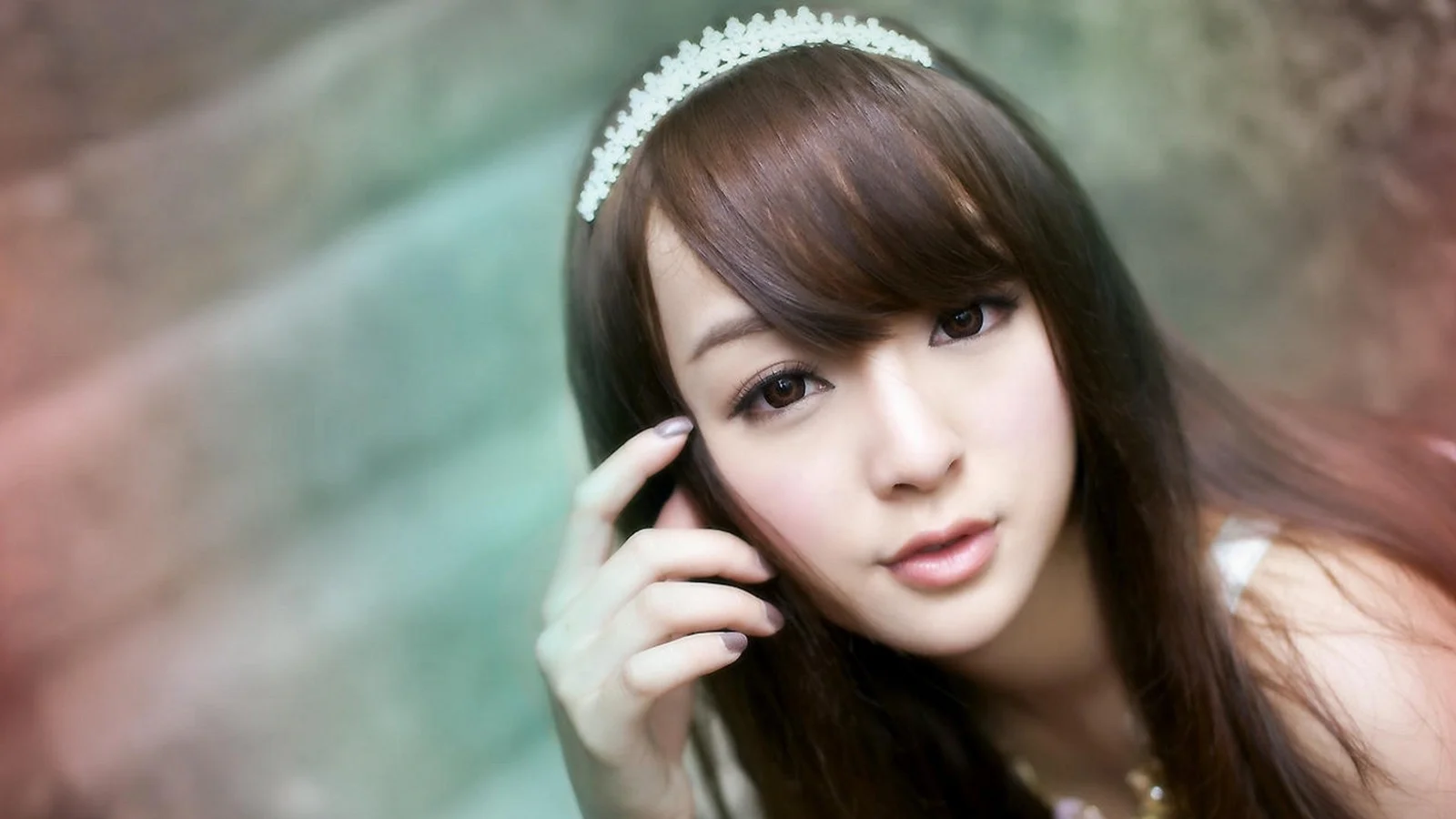 ティアラを身につけた茶髪の女性が、緑と茶色にぼかされた柔らかな背景の前で頬に指を添えながらカメラを見つめています。
