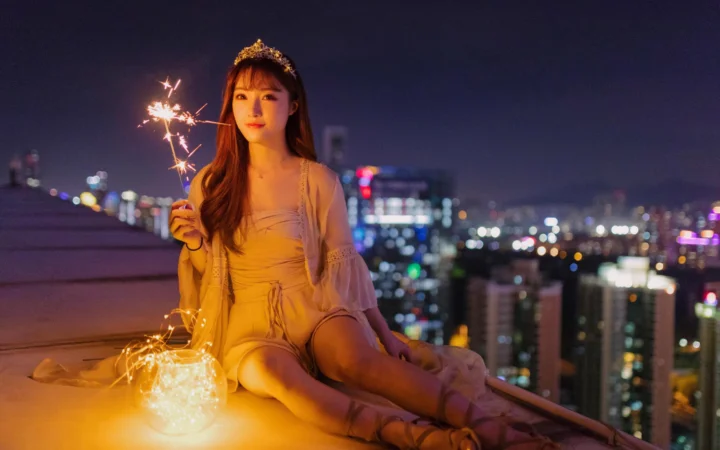 一名女性坐在屋顶上手持仙女棒，身后是朦胧的城市夜景。