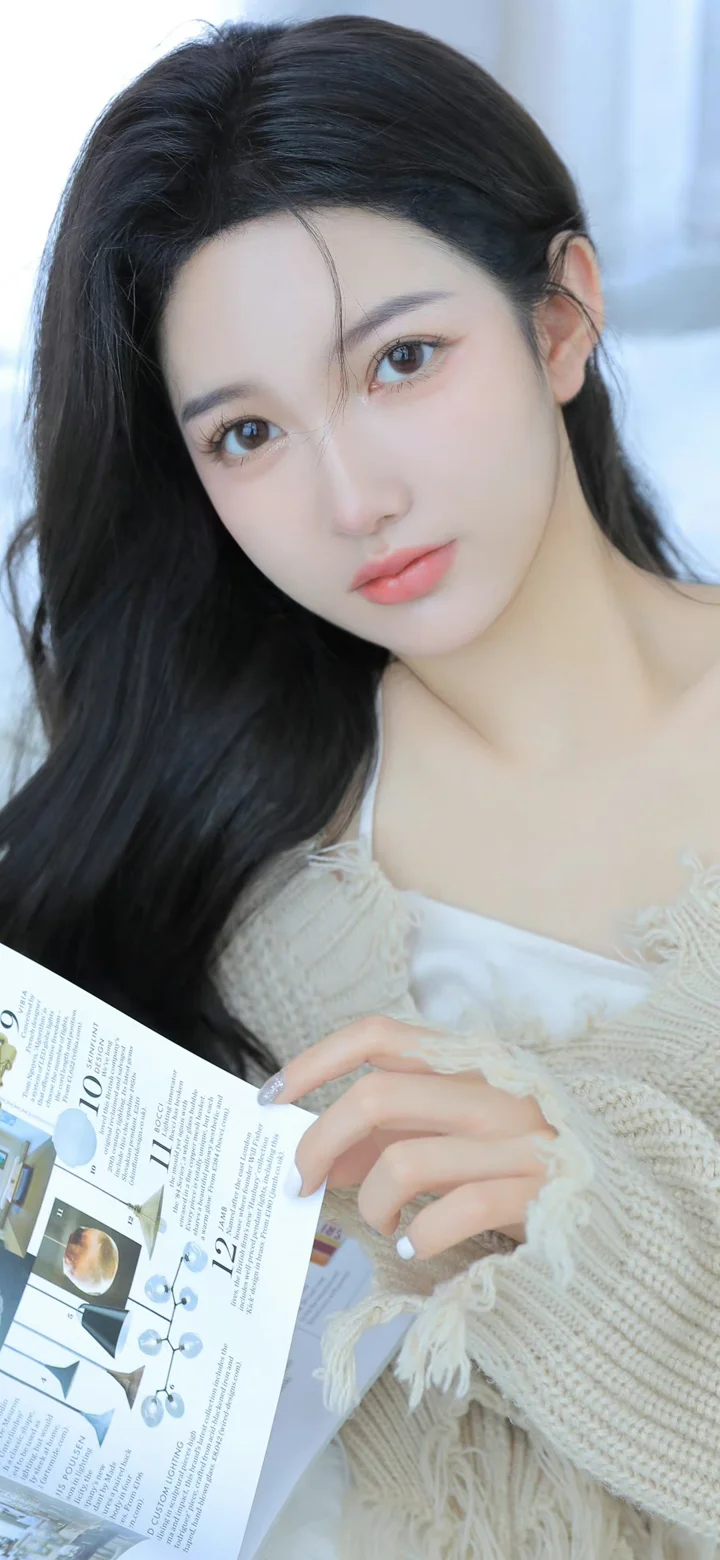 긴 머리의 여성이 밝은 색 니트를 입고 잡지를 들고 카메라를 바라보고 있습니다.