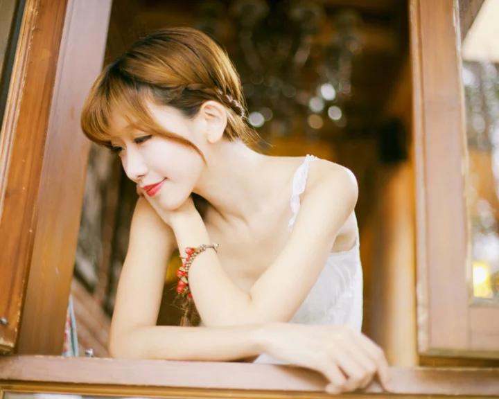 흰색 끈 민소매 상의를 입은 성인 여성이 나무 창가에 기대어 부드럽게 미소 짓고 있습니다.