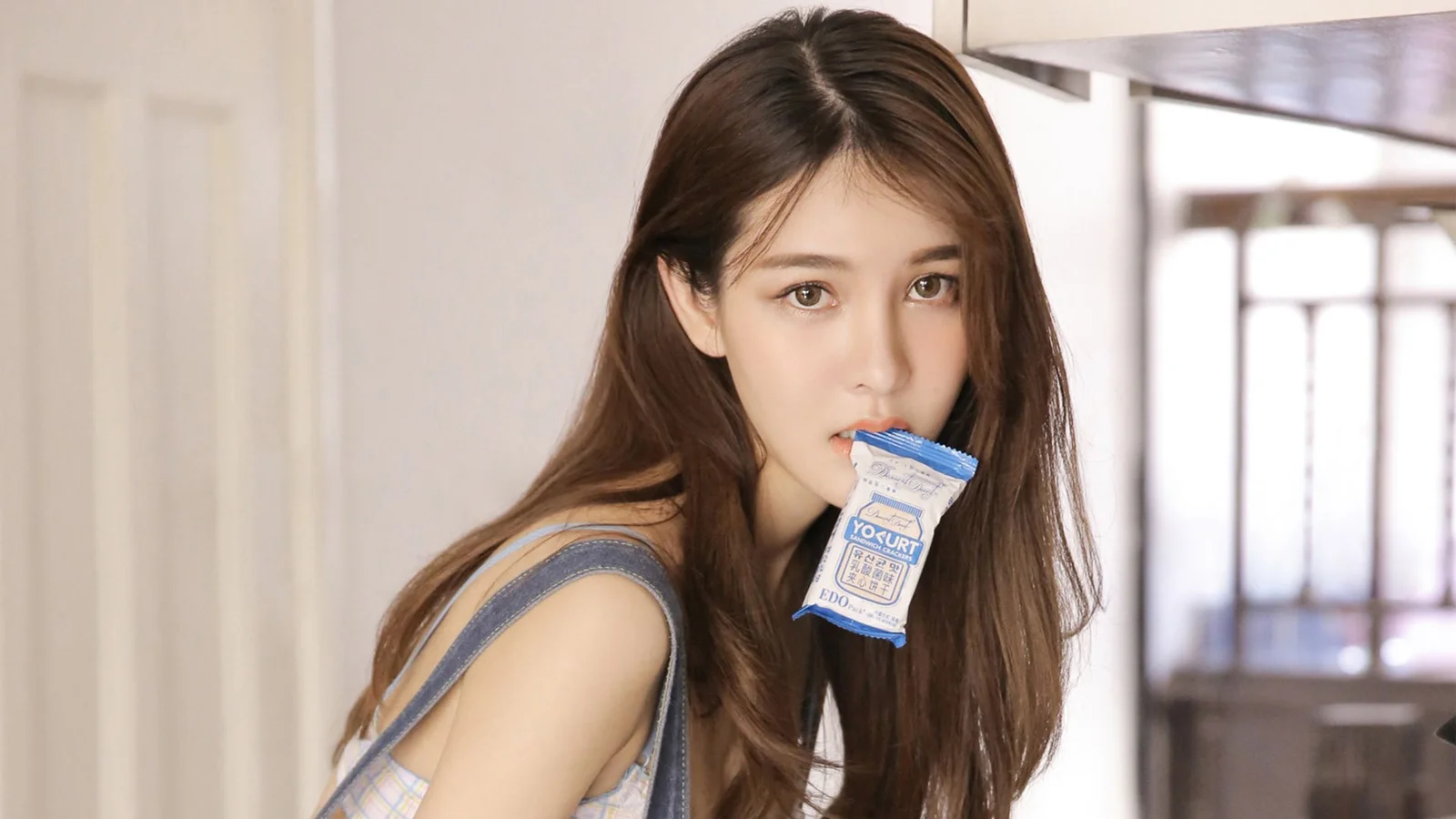 一名长发女子在室内靠近镜头，嘴边含着一小袋包装食品。