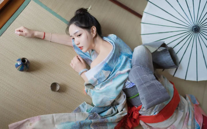 一名身穿淺藍色傳統服飾的女子躺在編織席上，旁邊放著一把白色紙傘。