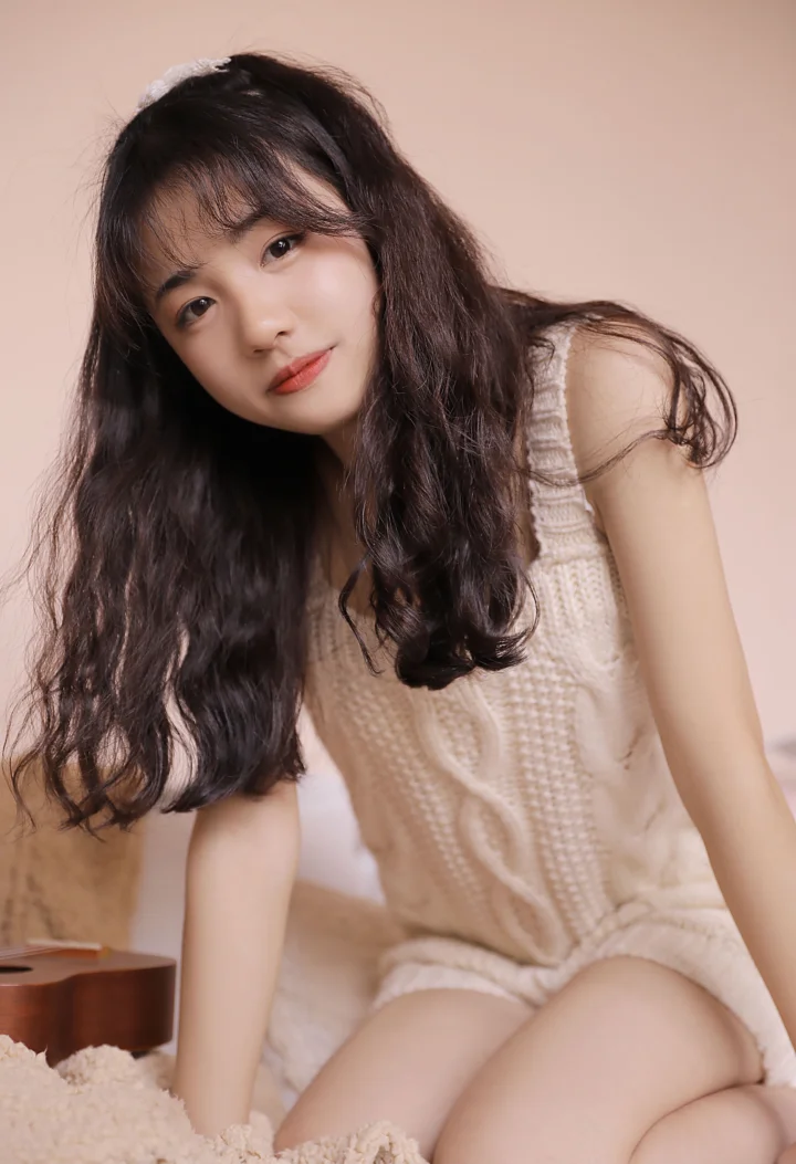 一名长发女性穿着浅色针织服装坐在室内柔和的粉色背景前。