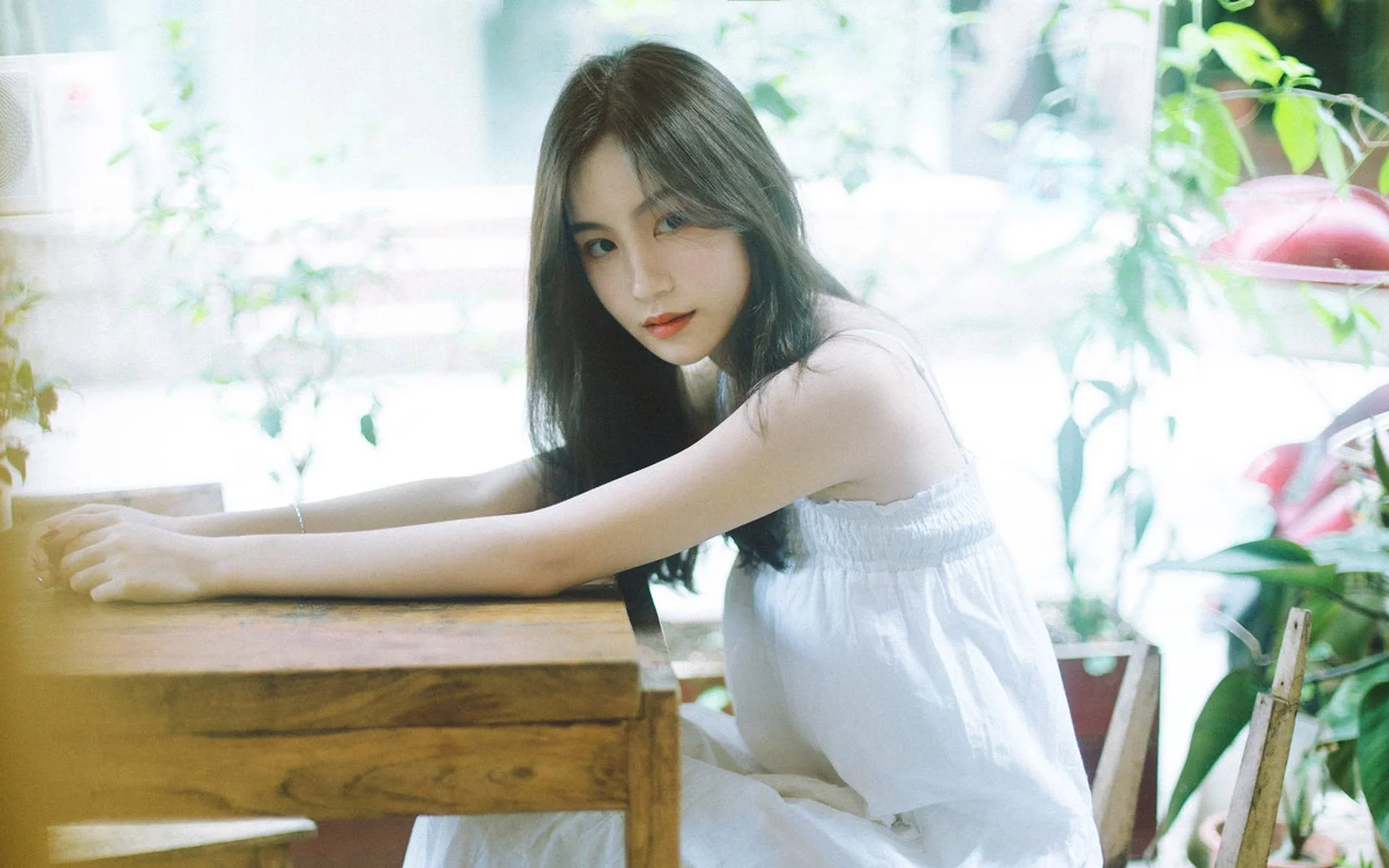 一名穿白色连衣裙的女子坐在木桌旁，室内窗边光线明亮。