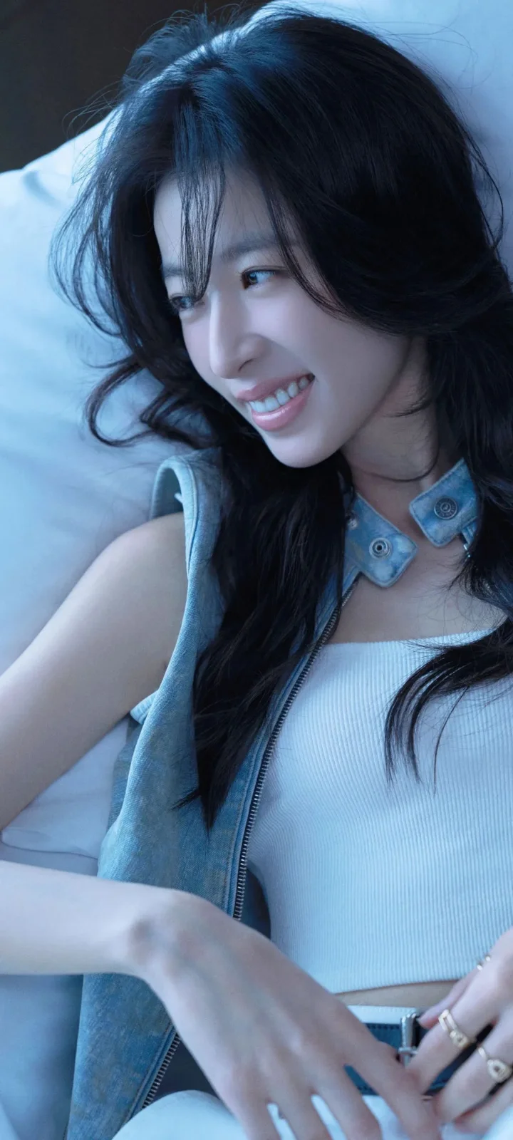 一名长黑发女性穿着牛仔背心和浅色上衣，在蓝色调光线下微笑。