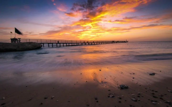 夕阳映照着海滩、海面和延伸入海的栈桥，天空与潮水呈现橙紫色光影。