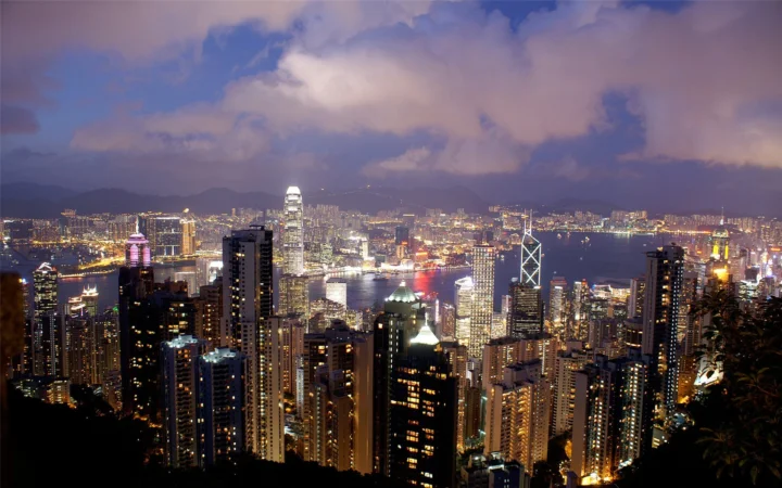 夜幕下灯火通明的高楼城市沿着港湾延伸，云层笼罩着远处天际线。