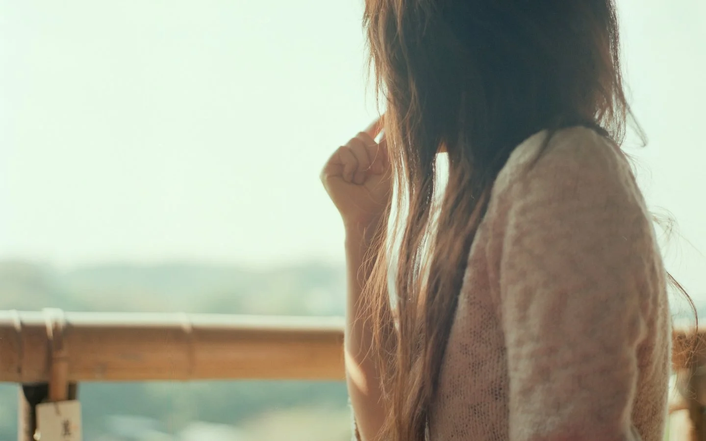 긴 머리의 여성이 밝은 야외 풍경을 바라보며 발코니에 서 있다.