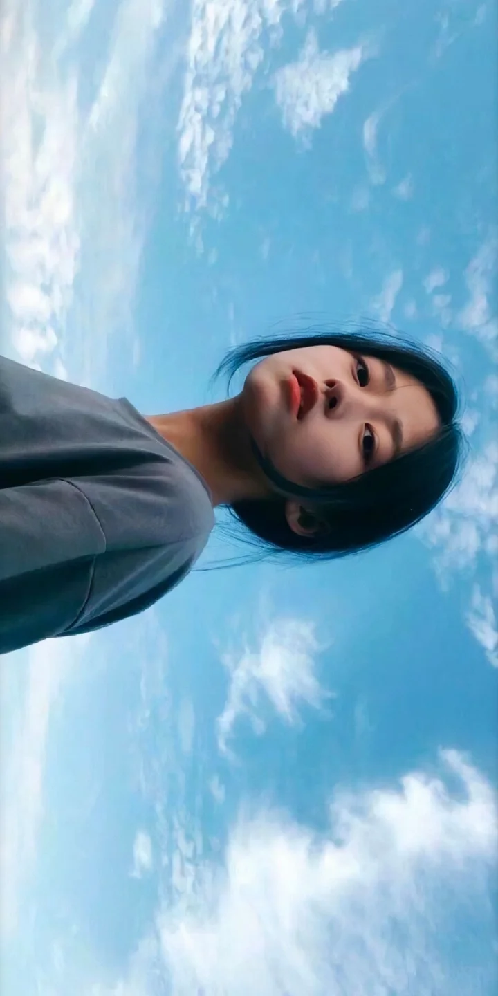회색 상의를 입은 여성이 푸른 하늘과 흰 구름 아래 서 있습니다.