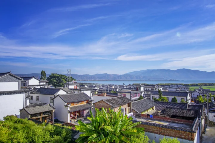 白族村落的传统屋顶延伸至洱海湖畔，远处是连绵山脉与蓝色天空。