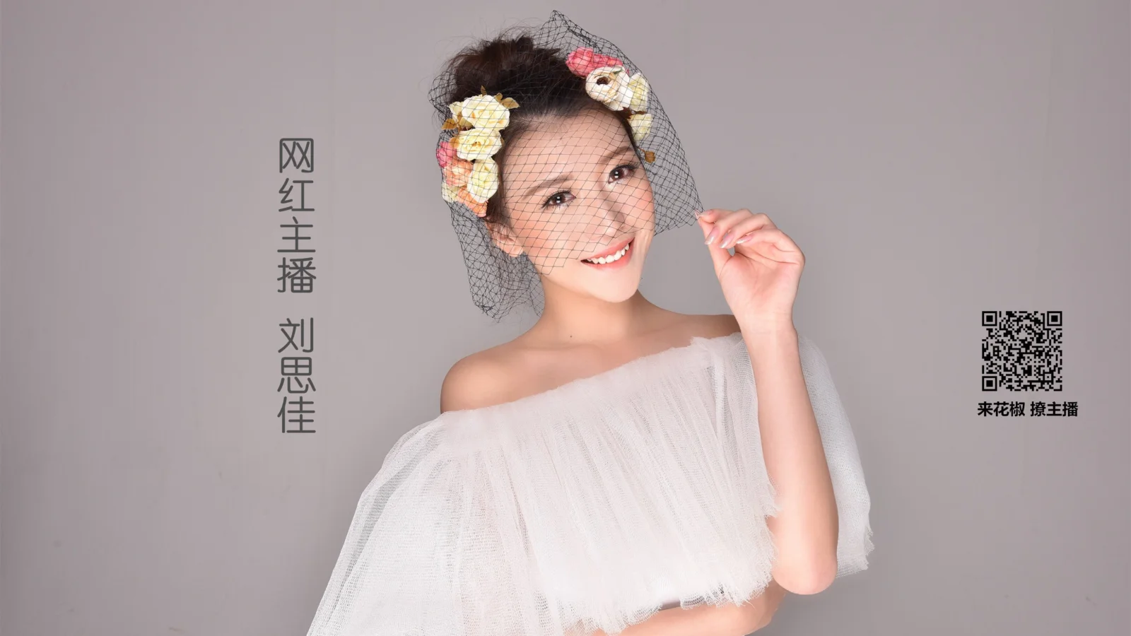 흰색 오프숄더 드레스를 입은 성인 여성이 꽃 장식과 베일을 착용하고 회색 배경 앞에서 미소를 지으며 포즈를 취하고 있습니다.