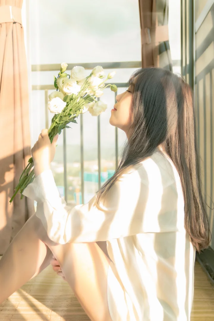 一名女子坐在窗边，手持一束白色鲜花，阳光洒在她身上。