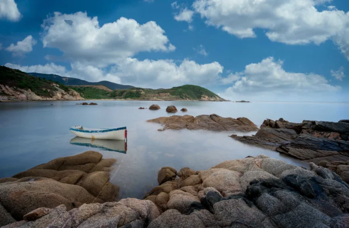 一艘小船停泊在宁静的海湾中，周围是岩石海岸、远山和蓝天白云。