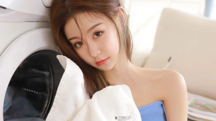 一名棕色长发女子穿着蓝色上衣，手持白色毛巾靠在洗衣机旁。