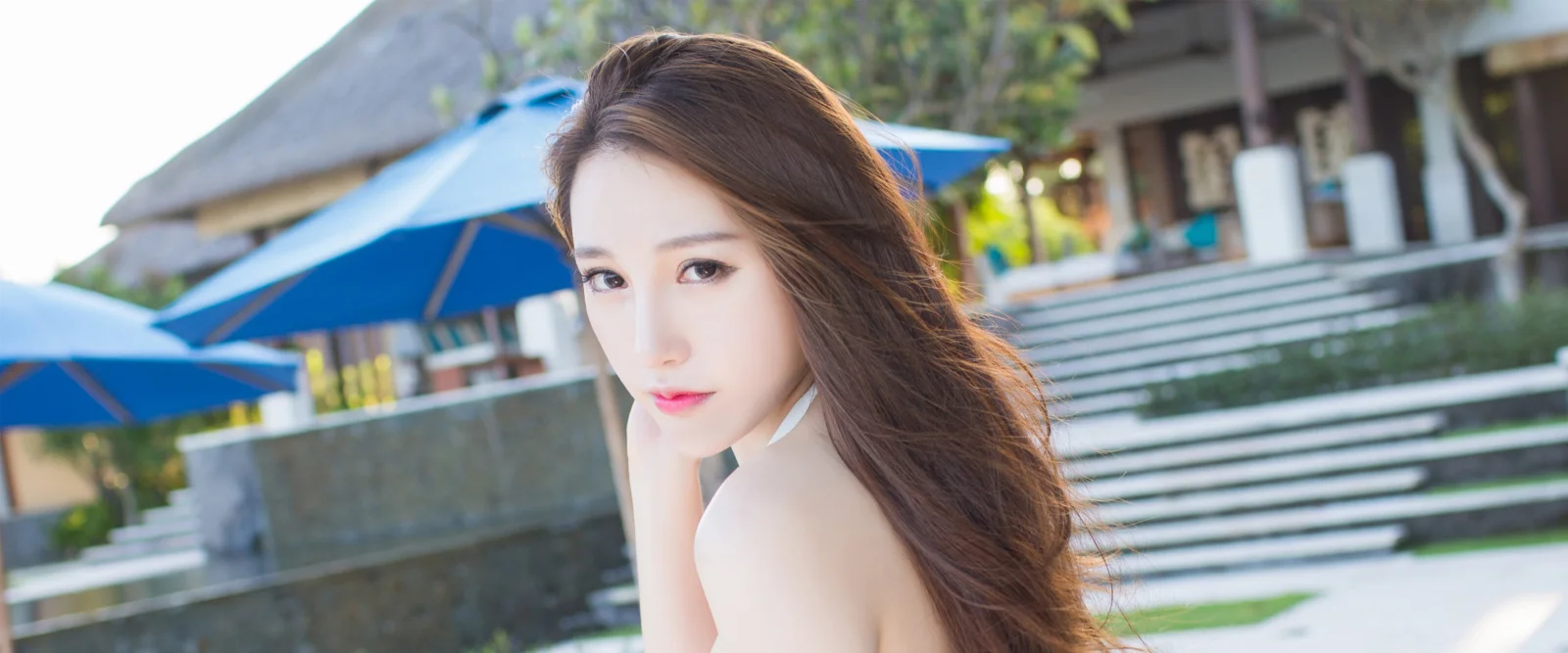 一位成年女性在户外度假村环境中回望镜头，背景可见蓝色遮阳伞和建筑台阶。