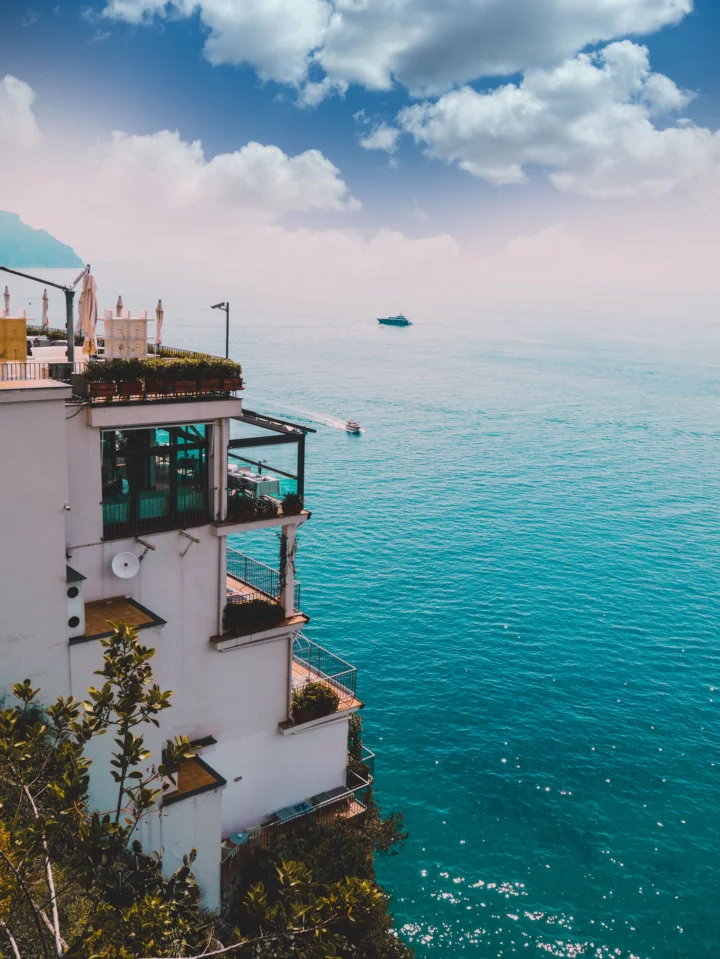 临海建筑坐落在画面左侧，远处是开阔的 turquoise 海面与漂浮的白云。