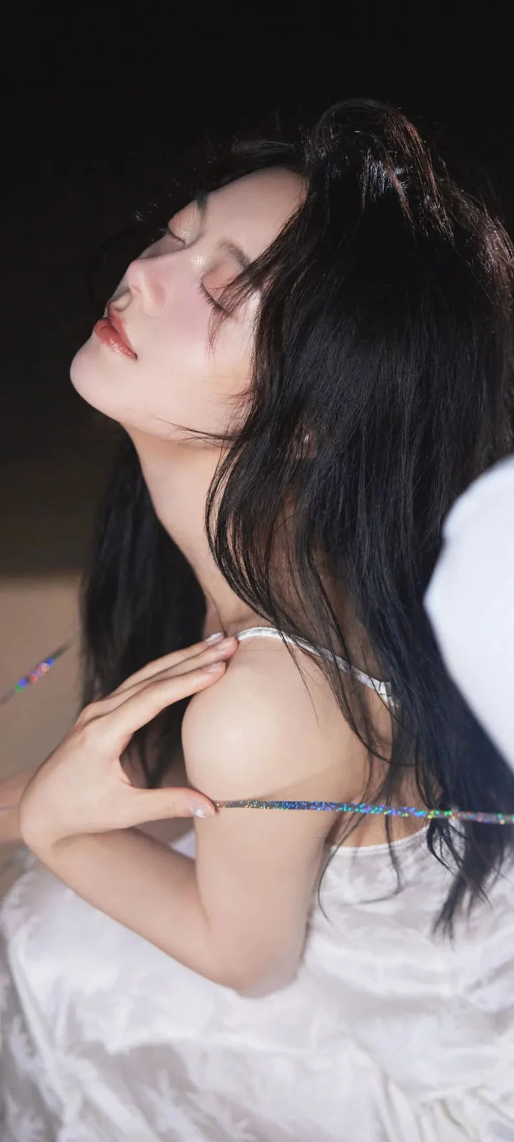 一位长发女子闭眼侧脸靠在深色背景前，身穿白色服装。