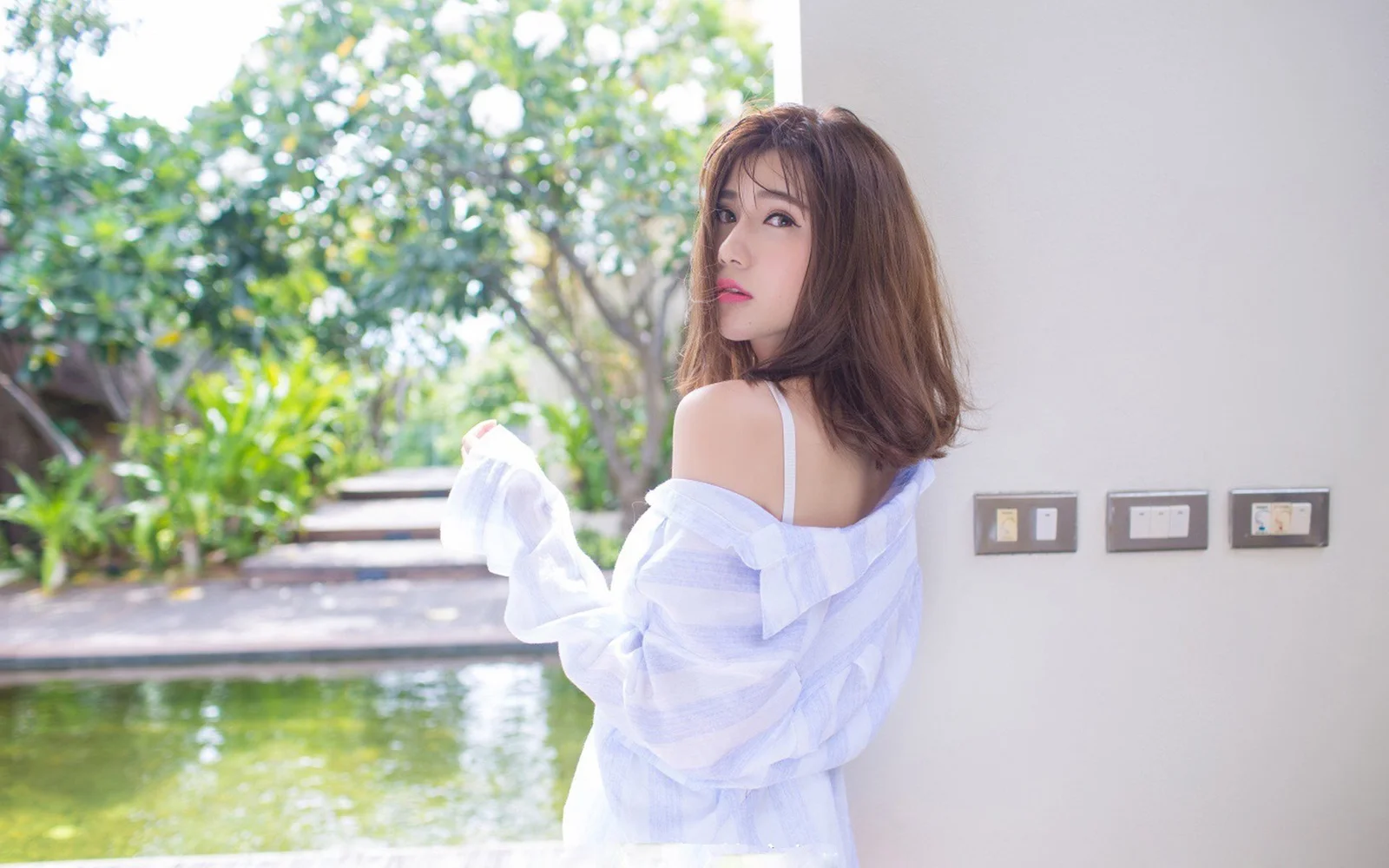 一名穿白色露肩上衣的女子站在花园泳池旁回望镜头。