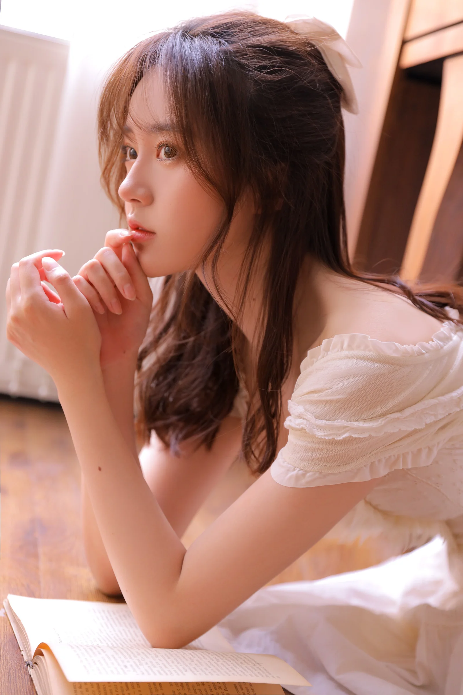 一名长发女性穿着白色上衣，俯身靠在书本旁望向窗边。