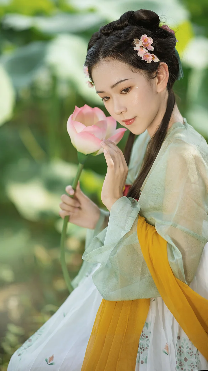 一位身穿传统服饰的女子在绿色户外环境中手持粉色荷花。