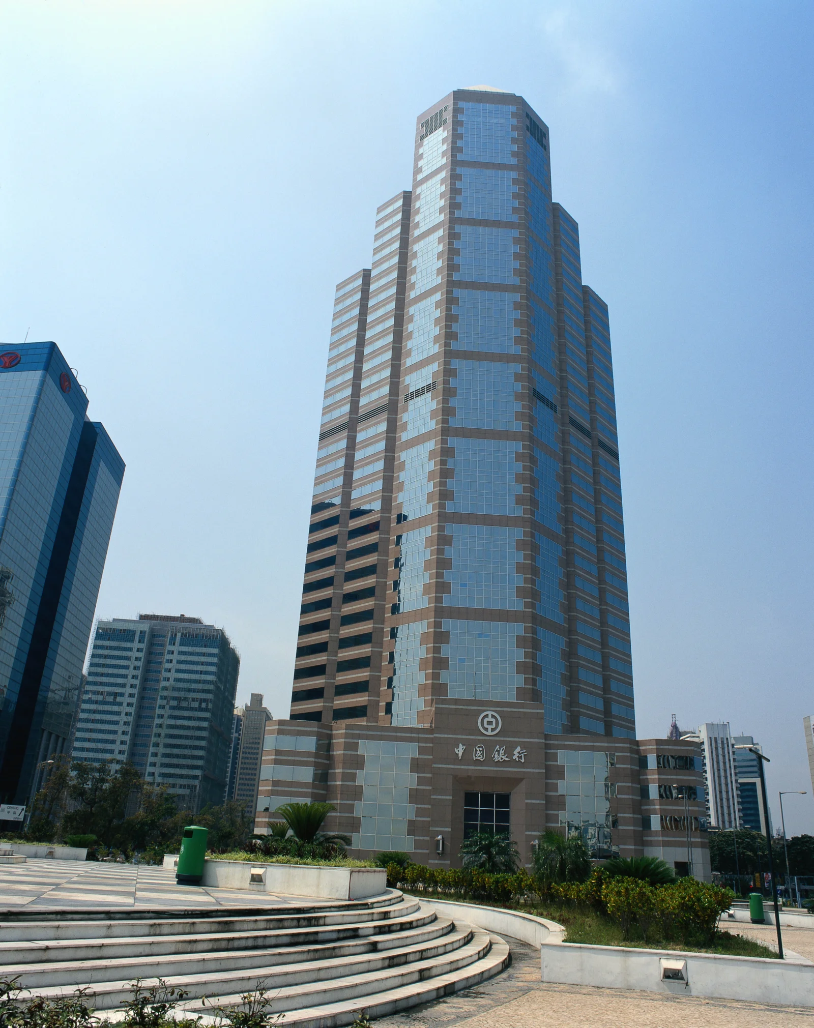 蓝天下，一座现代高层建筑矗立在城市广场旁，周围可见其他办公楼和阶梯景观。