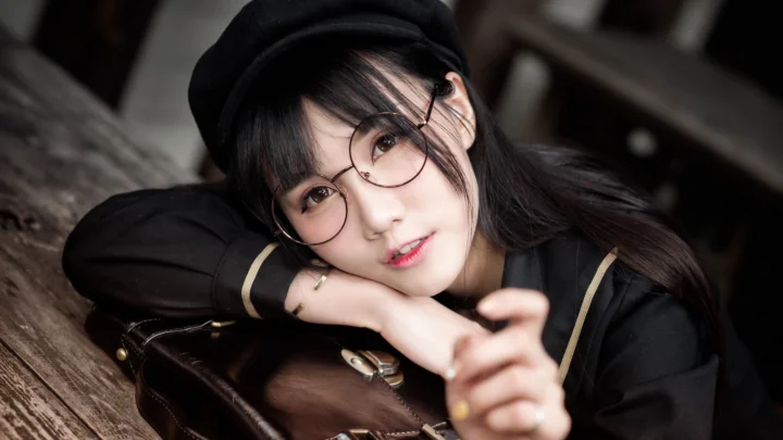 一名戴圓框眼鏡和黑色帽子的女性靠在木桌旁看向鏡頭。