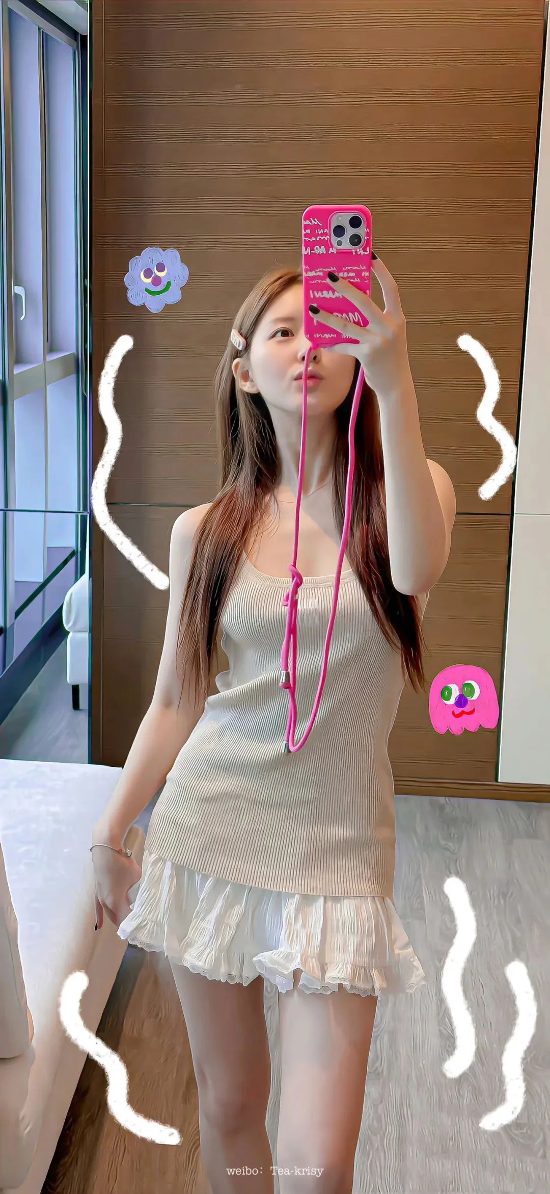 成人女性が室内の鏡の前でピンク色のスマートフォンを持って自撮りしており、淡い色のトップスとフリル付きの短いスカートを着ている。