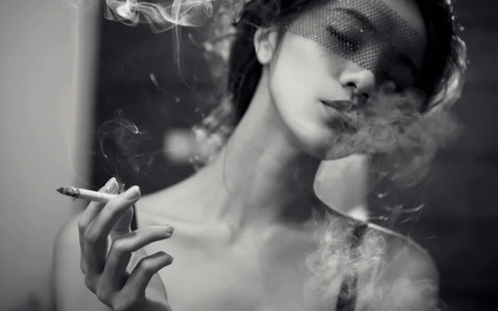 黑白画面中，一名戴着面纱的女性手持香烟，周围缭绕着烟雾。
