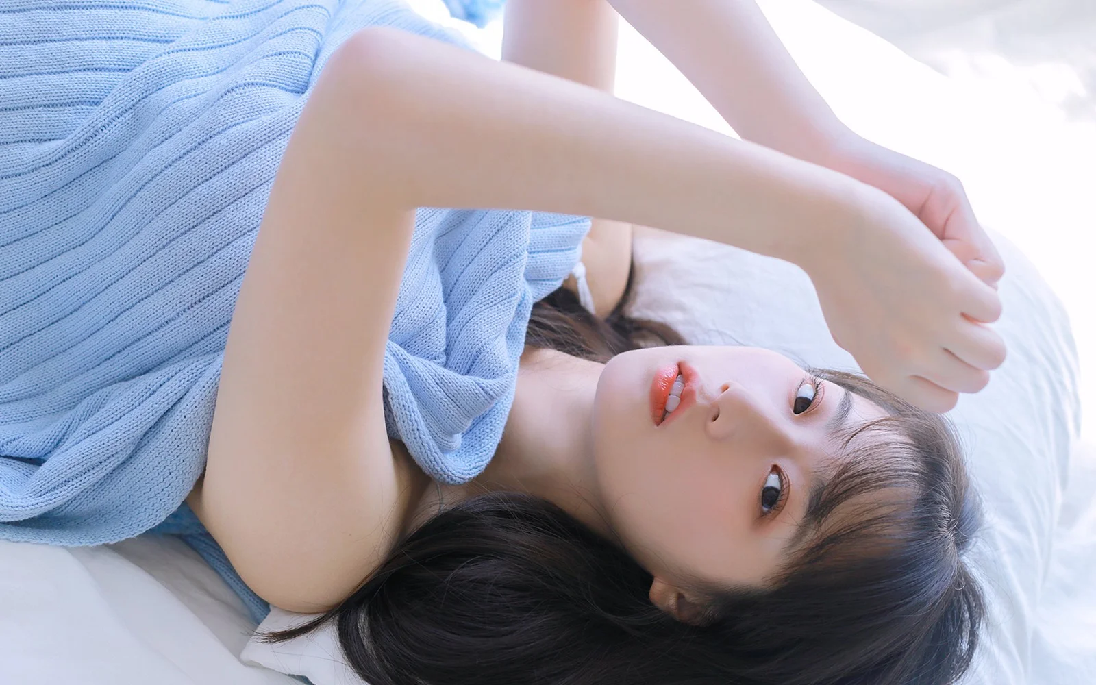 연한 파란색 니트 스웨터를 입은 성인 여성이 침대에 누워 카메라를 바라보고 있습니다.