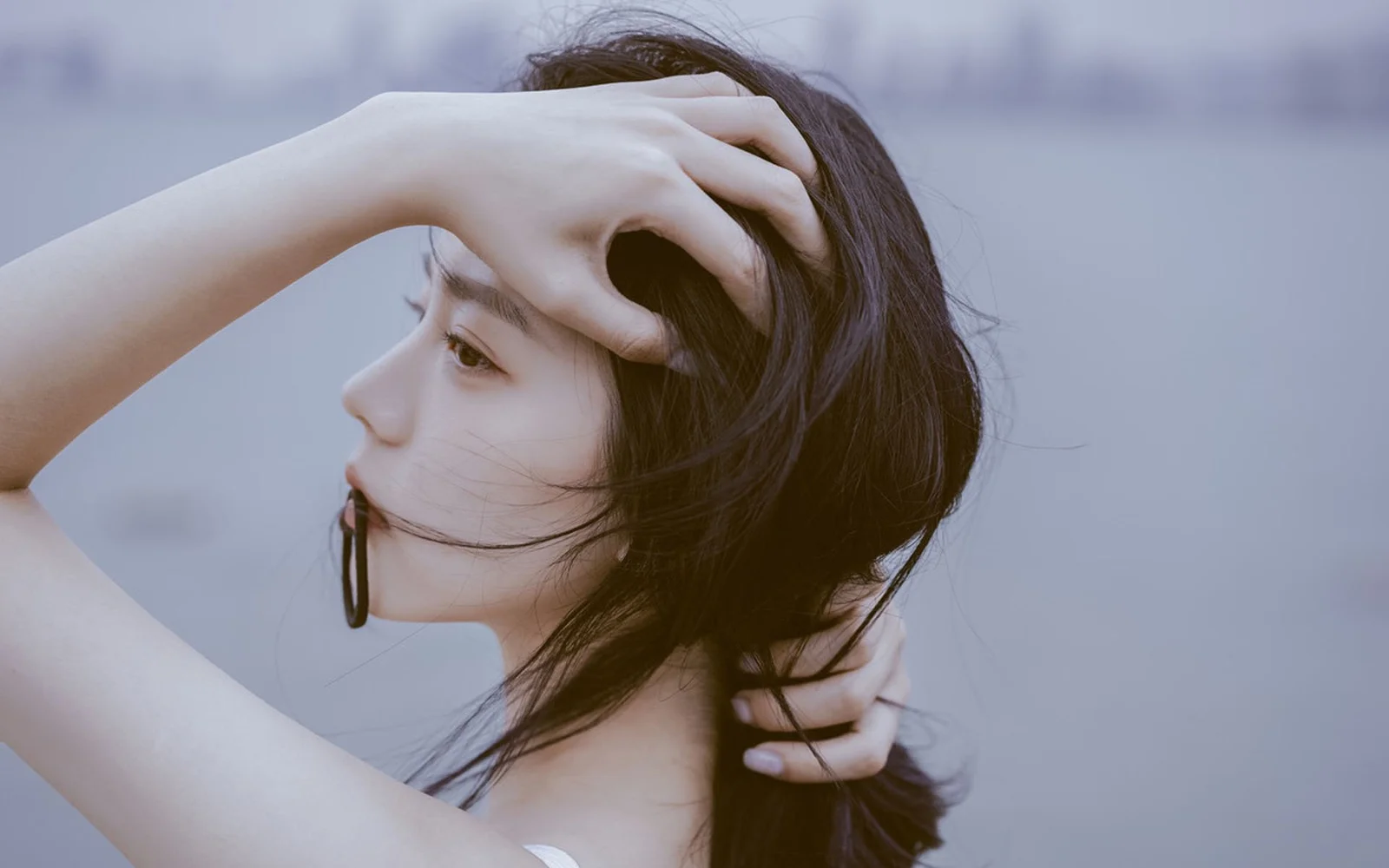 一名黑发女性侧脸近距离出现在柔和的蓝灰色背景前，双手整理着头发。