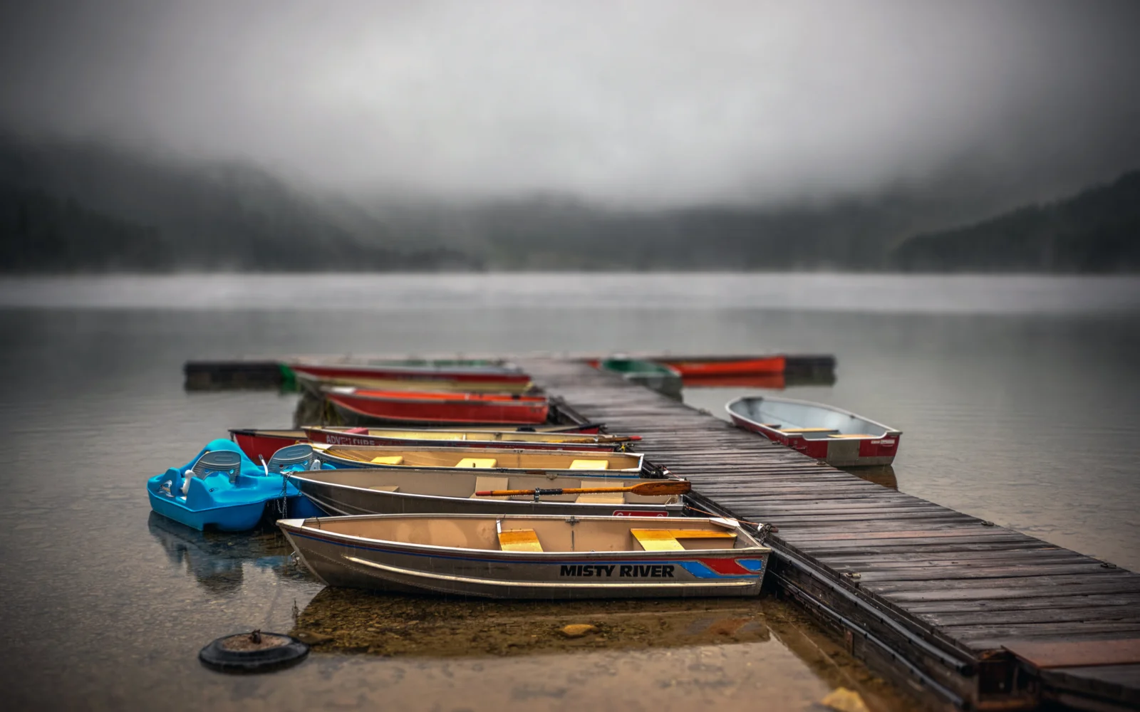A row of small boats rests beside a wooden dock on a misty lake, with hazy forested hills in the distance.
