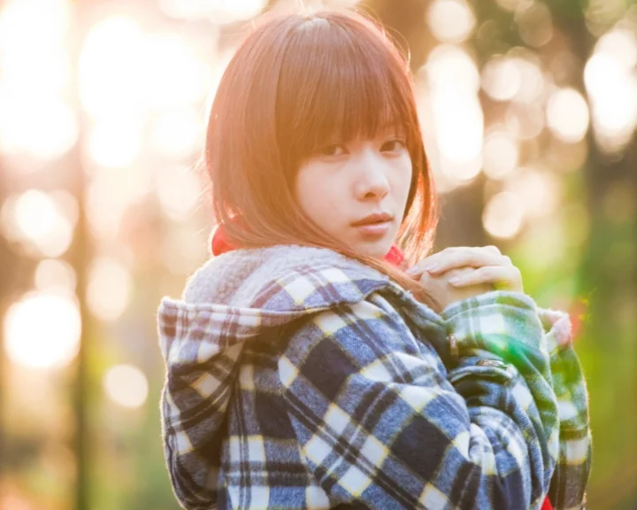 一名留着齐刘海的女子穿着格纹外套站在阳光斑驳的树林中回望镜头。