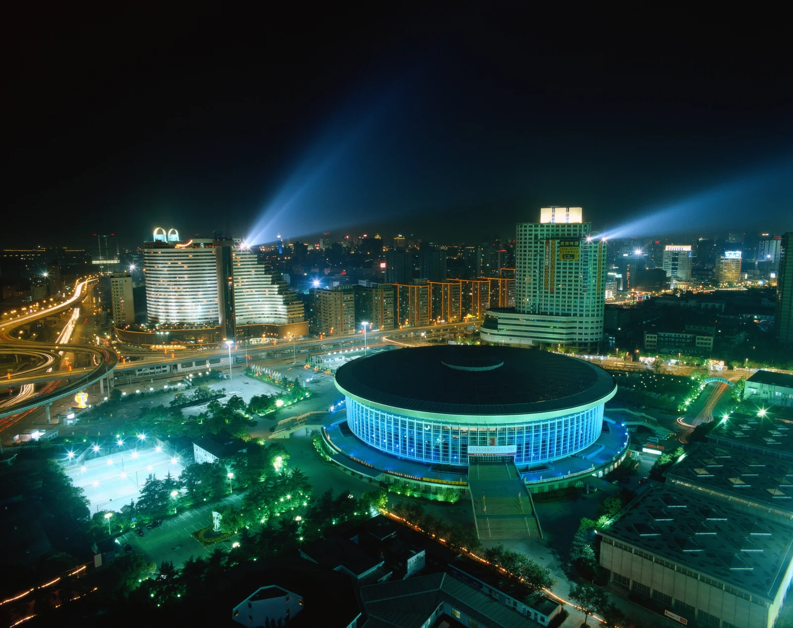 夜色中的城市建筑与道路灯光环绕着一座大型椭圆形场馆，构成明亮的城市夜景。