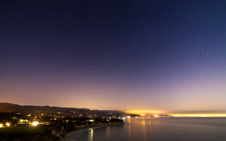 繁星点缀的夜空下，海湾、山丘和沿岸灯光倒映在平静的海面上。