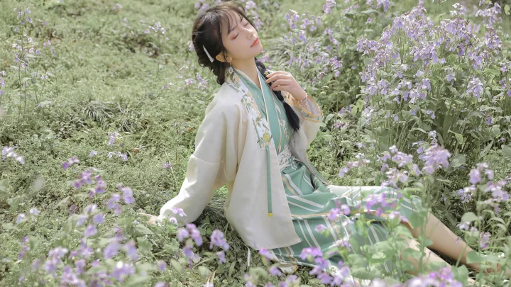 한 여성이 밝은 색상의 옷을 입고 보라색 들꽃 사이에 앉아 있으며 주변은 푸른 식물로 둘러싸여 있습니다.