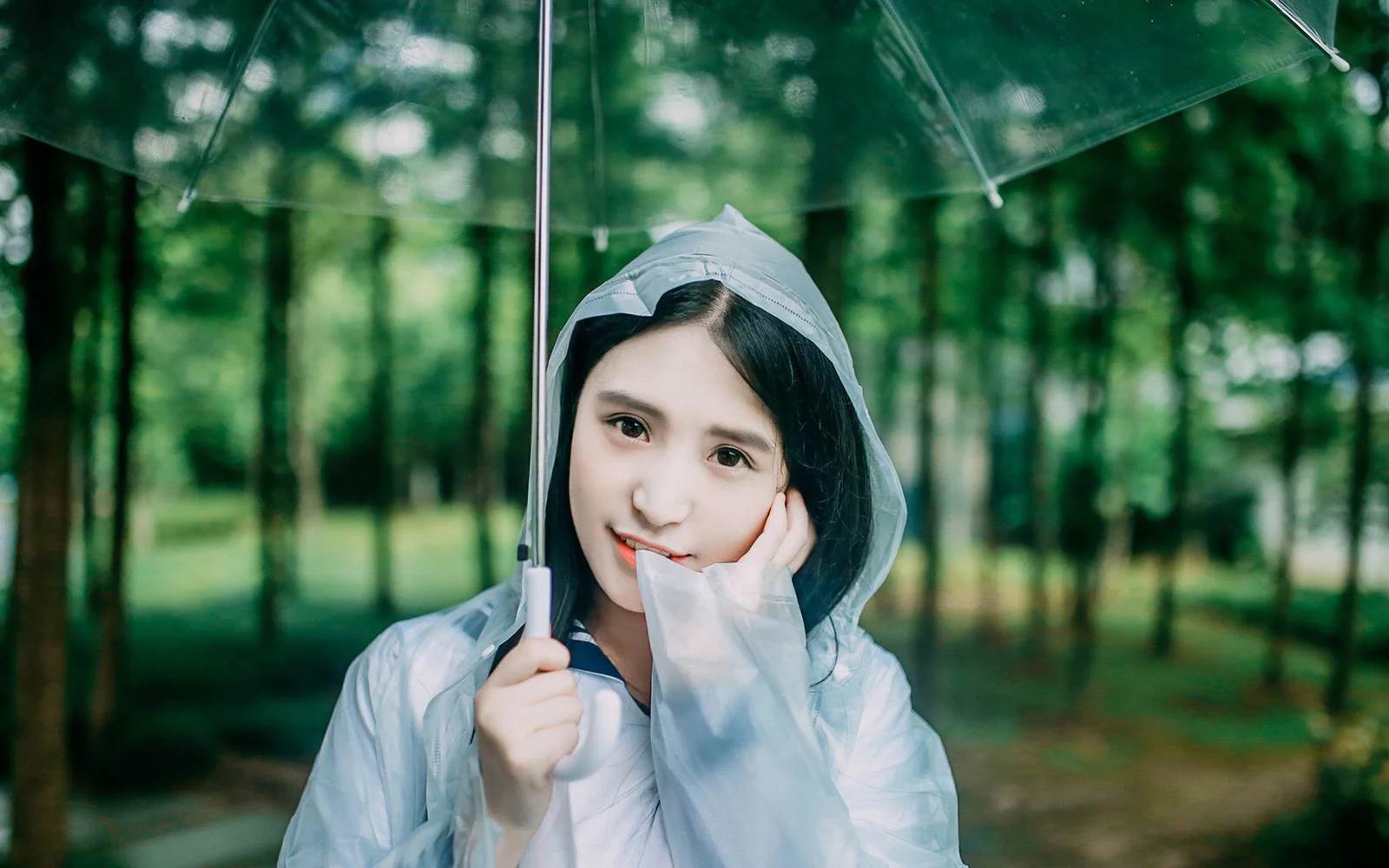 一名穿著透明雨衣的年輕女子撐傘站在綠色森林中。