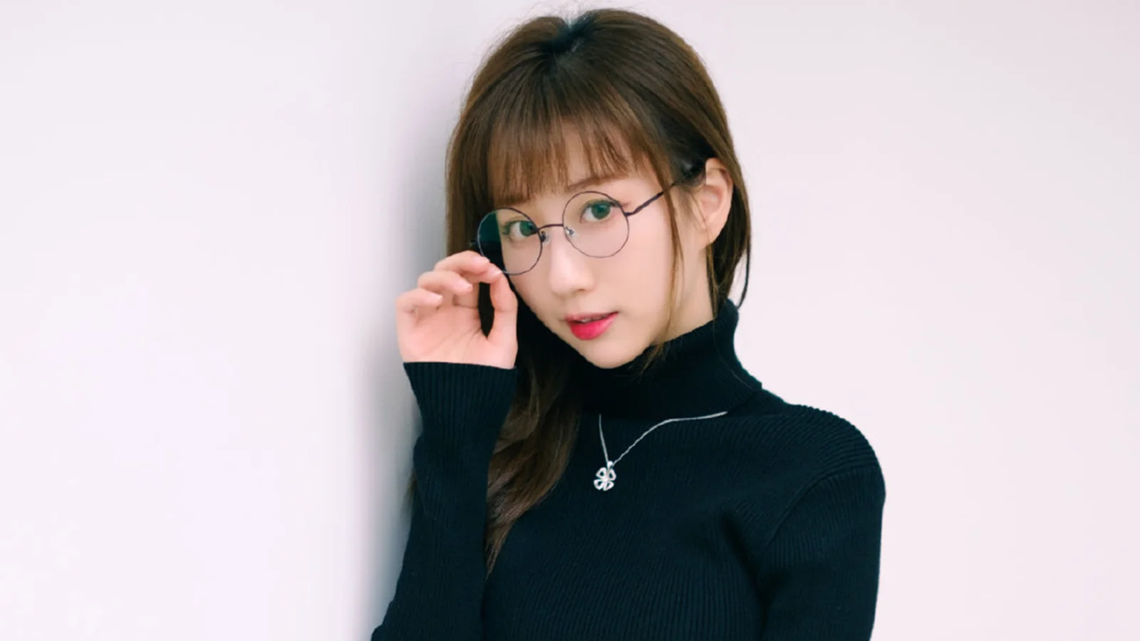 一位戴圆框眼镜、穿黑色高领毛衣的女性靠墙注视镜头。