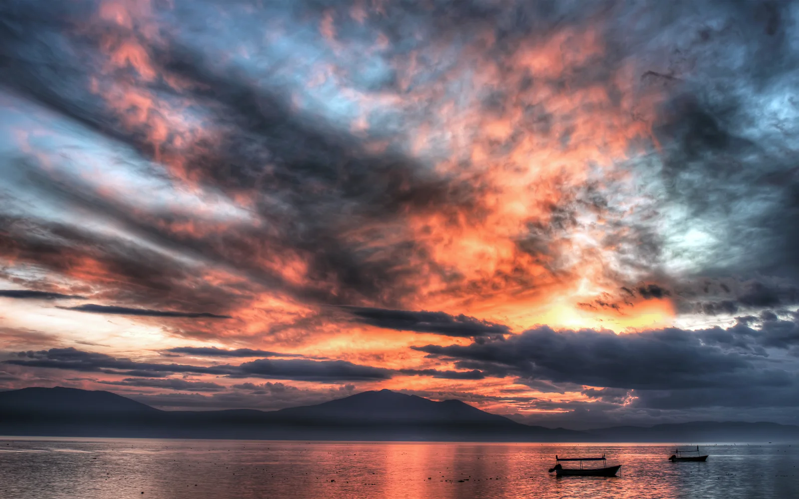 鮮やかな橙色と赤色の夕焼け雲が、遠くの山々と穏やかな湖面、二隻の小舟を照らしている。