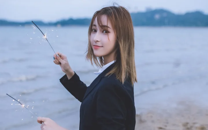 一名女子站在海邊，手持點燃的仙女棒，背景是平靜的海面與遠處的海岸線。
