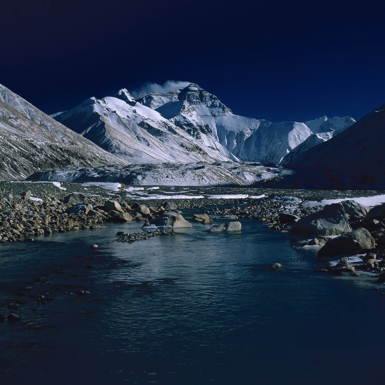 雪山与冰川映衬着深蓝天空，前景是一条流经岩石的河流。